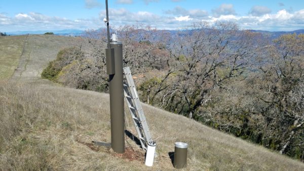 Tall metal rain gauge with metal ladder leaning against it on grassy hillside with oaks and mountains in background.