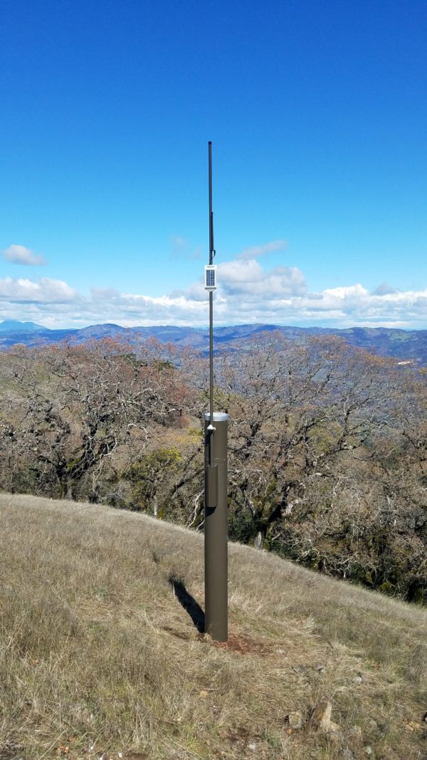 Vertical shot of tall metal rain gauge on grassy hillside with oaks in the background and mountains in the distance.