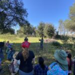 A crowd of community members stand in the grass and listen to speakers share about the Dogbane plant.