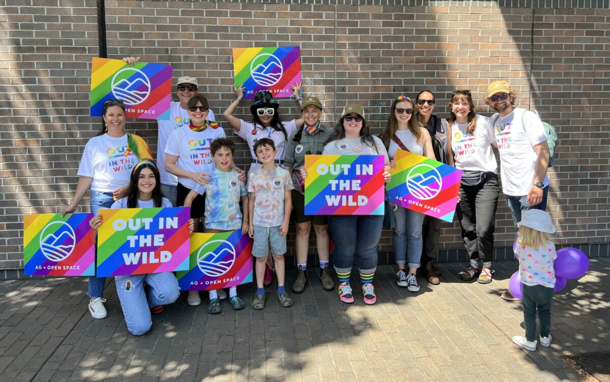 Ag + Open Space staff pose at the Sonoma County Pride festival. They are smiling at the camera, holding signs with rainbow background that say Out in the Wild and have the Ag + Open Space logo.