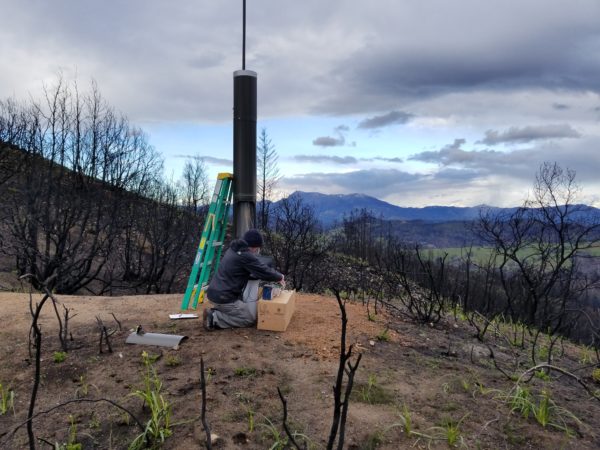 A man installs a 10-12 foot rainfall gauge on a burned hillside with charred trees.