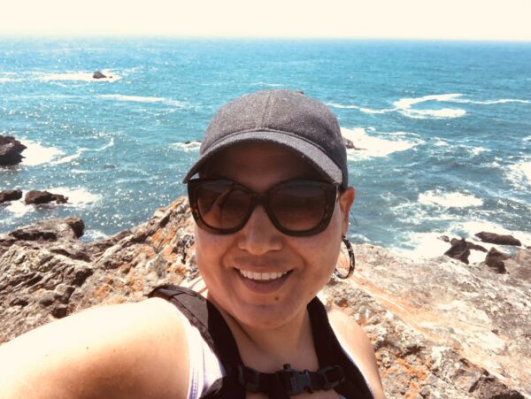A selfie of Irma Cuevas, where she is standing in front of coastal rocks and blue Pacific Ocean. She is wearing a black baseball camp, and large dark sunglasses, and smiling at the camera. 