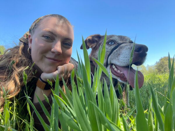 Kendall looking at the camera, while resting her chin on her palm, while lying in the grass next to her dog. 