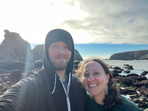 Selfie de Lauren y su esposo, mirando a la cámara con rocas costeras y agua de fondo. 