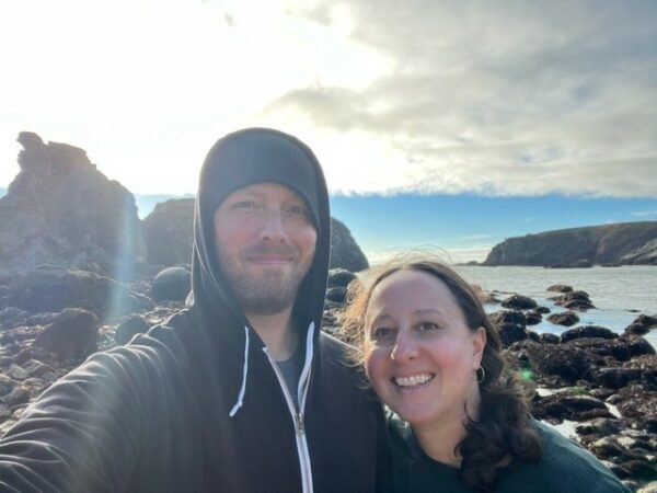 Selfie of Lauren and her husband, looking at the camera with coastal rocks and water in background. 