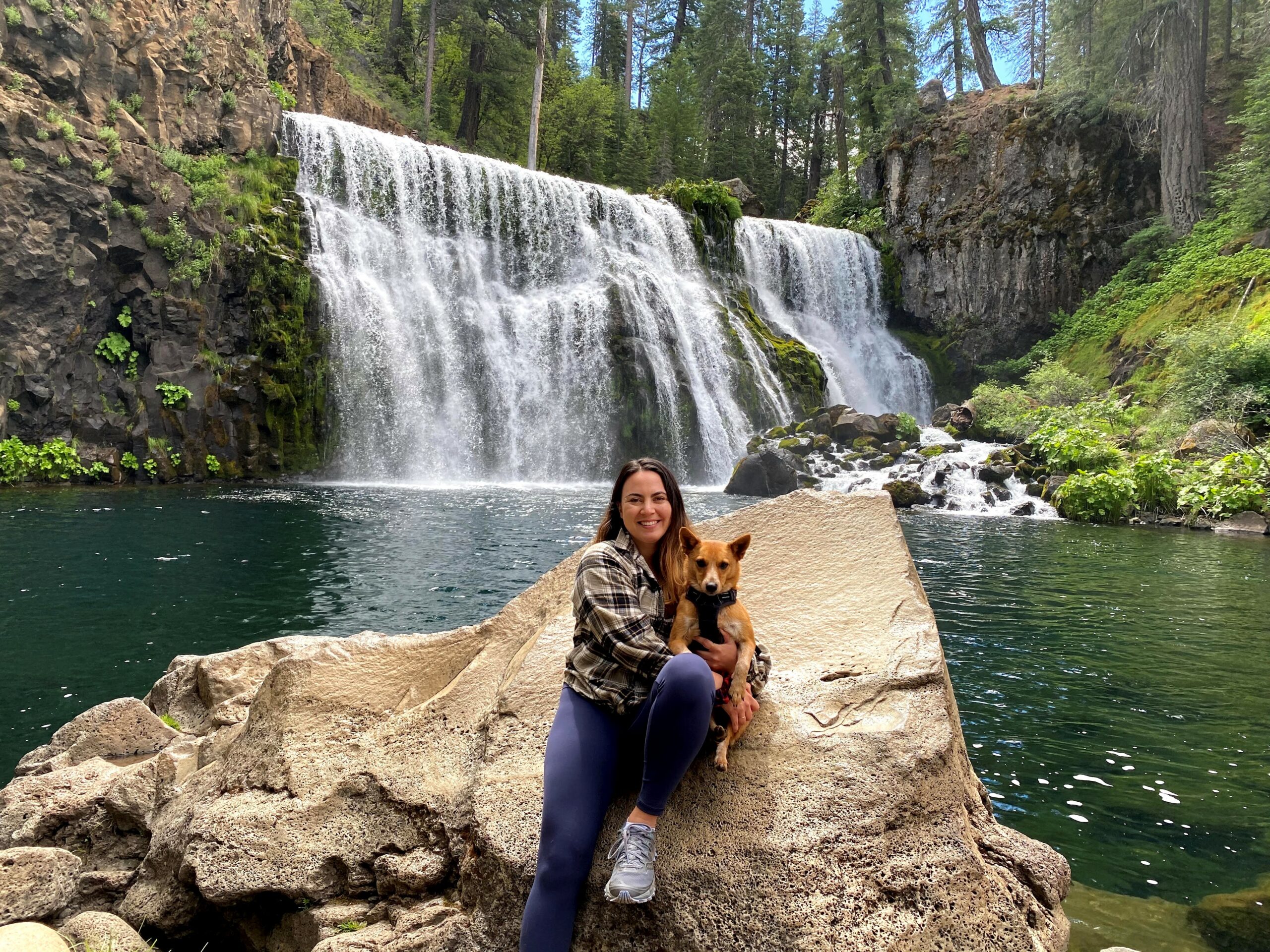 Marina holding her dog while sitting on a large rock in front of a waterfall.