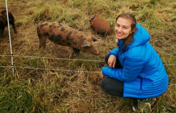 Mary Chambers crouched in a field, posing near pigs while smiling up at the camera.