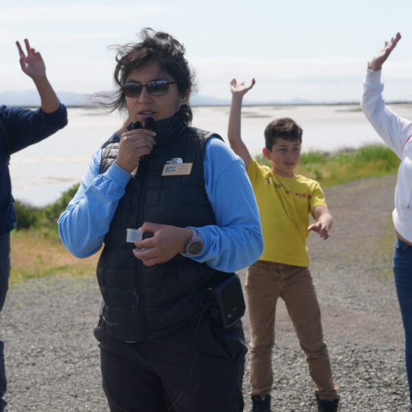 Photo of Mirella Ramos, standing outside with people behind her raising their hands. Mirella is wearing a blue shirt with a black vest, and they are standing in front of a body of water.