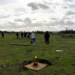 Students walking away across a green field, with a small area of cleared earth marked with a flag where a native plant has been planted.