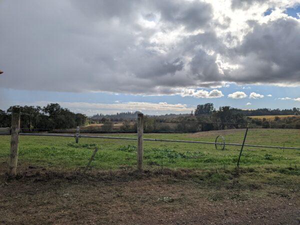 Landscape view of wooden fence, green field, and trees and hills in the distance. The sky is blue but has large white and grey clouds.
