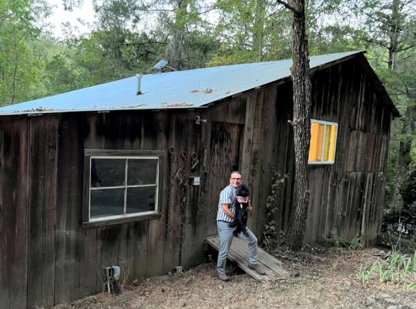 Photo of Pierre, standing front of a wood building, holding his dog Sydney in his arms. 