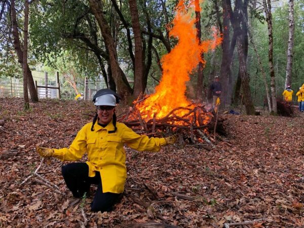 Raquel kneeling in front of a pile of branches on fire during a prescribed burn. Raquel is facing the camera with her arms held wide, and wearing a yellow safety jackets and a white hard hat.