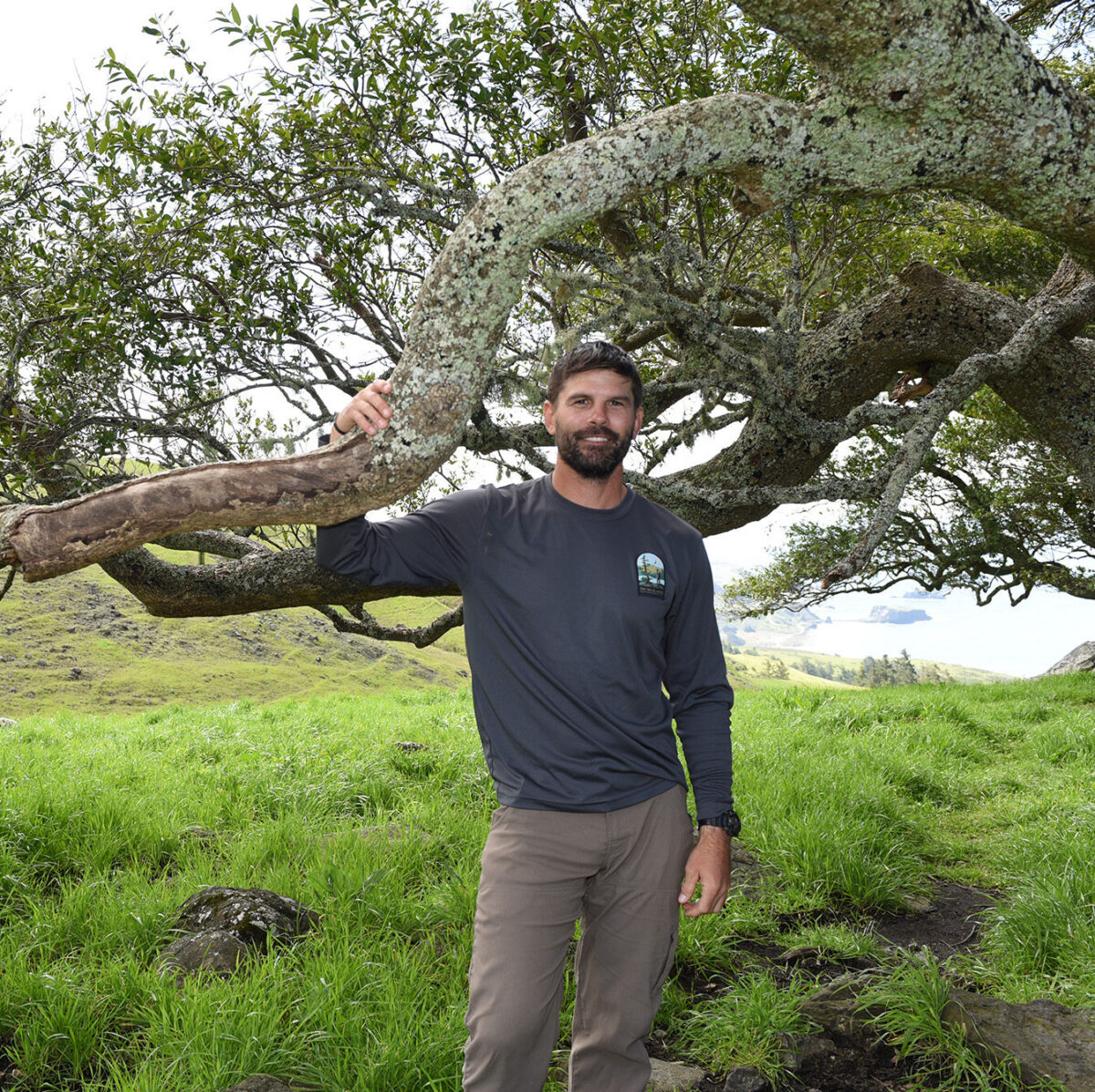 Photo of Ryan Berger, wearing a black shirt and brown pants. He's smiling at the camera while having one hand holding on to a large tree branch.