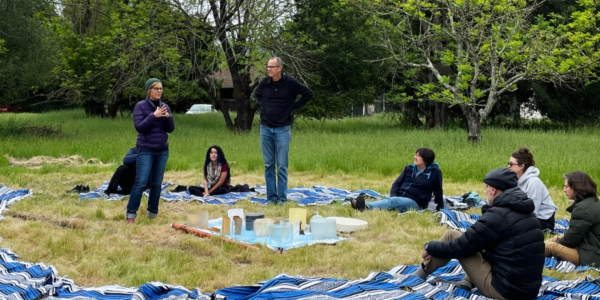 People sitting in a circle on blankets outside on grass. Two people in center talking to hem.