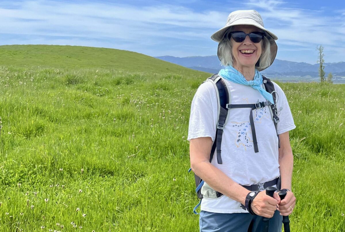 Photo of Wendy Eliot standing in a green field. She is wearing a tan hat, sunglasses, a blue scarf, white shirt, backpack, and using hiking poles. She's smiling at the camera.
